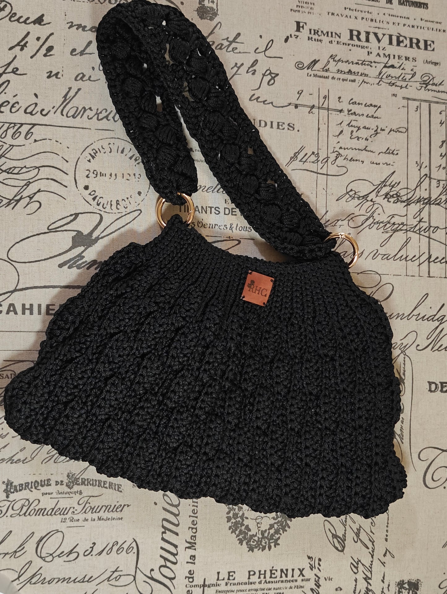 A black crocheted shoulder bag with a wide strap and gold-tone hardware, laid flat on a vintage-style printed background.