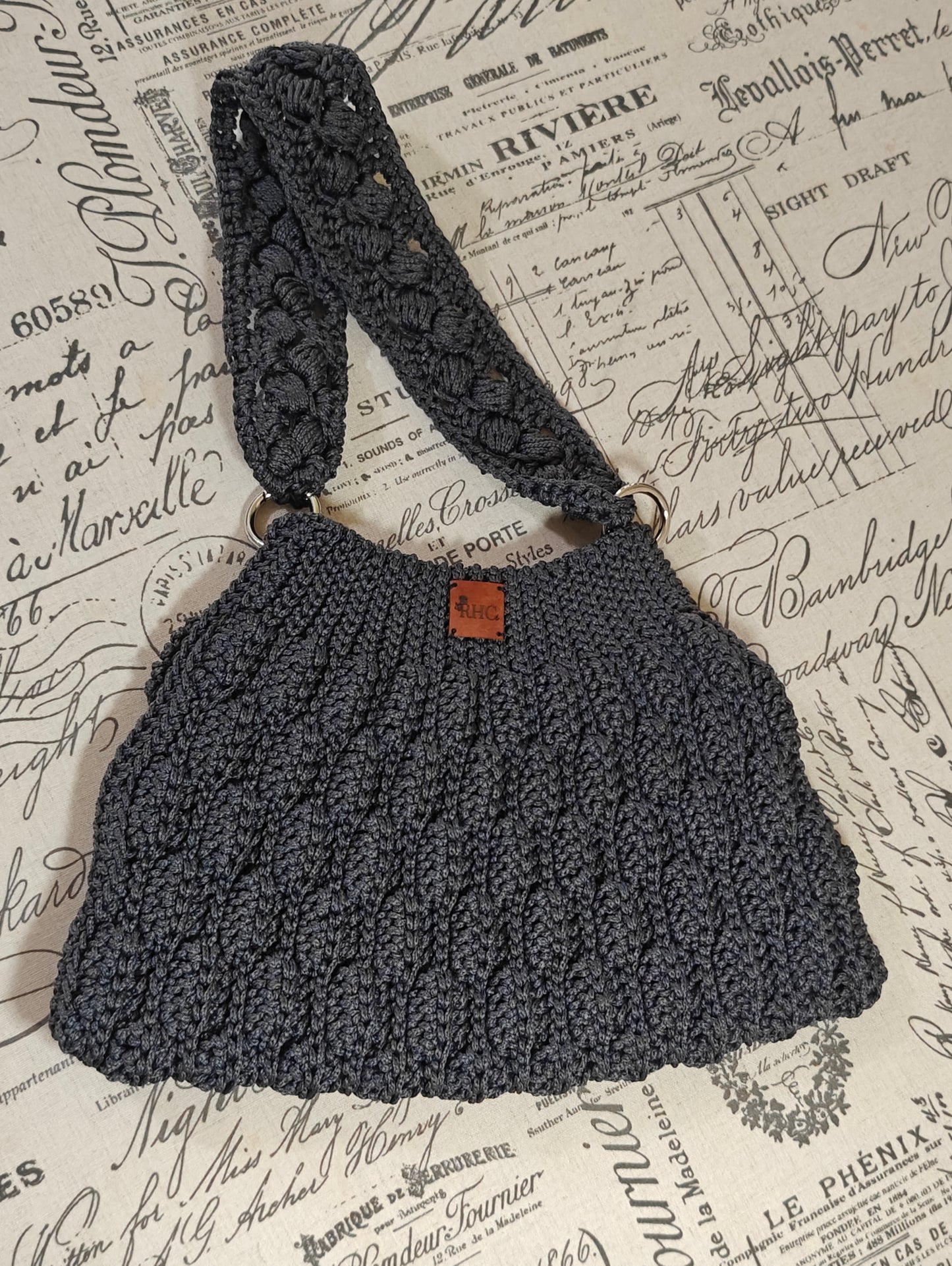 A dark gray crocheted shoulder bag with a wide strap and a small brown leather brand patch, displayed on a vintage-style script paper background.