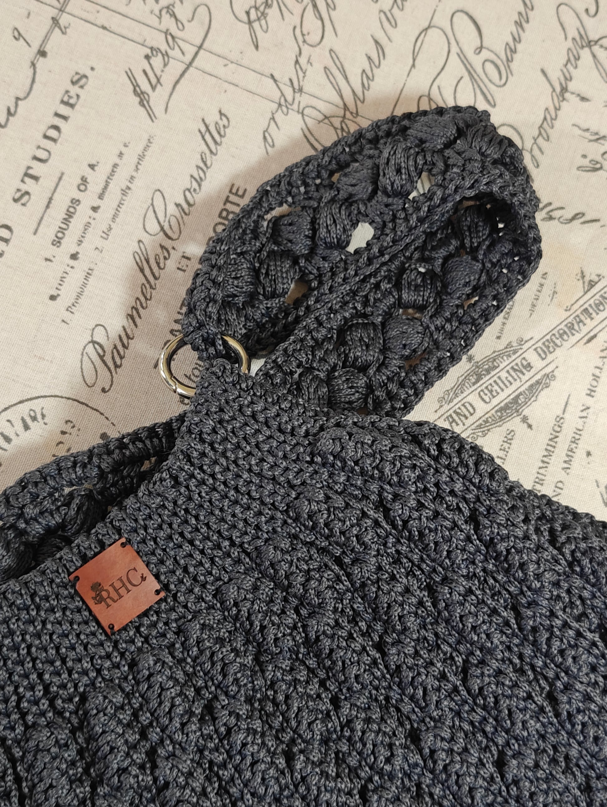 Close-up of a dark charcoal gray crocheted handbag with a textured shoulder strap and a small brown leather 'RHC' brand patch, laid flat on vintage-style printed paper.