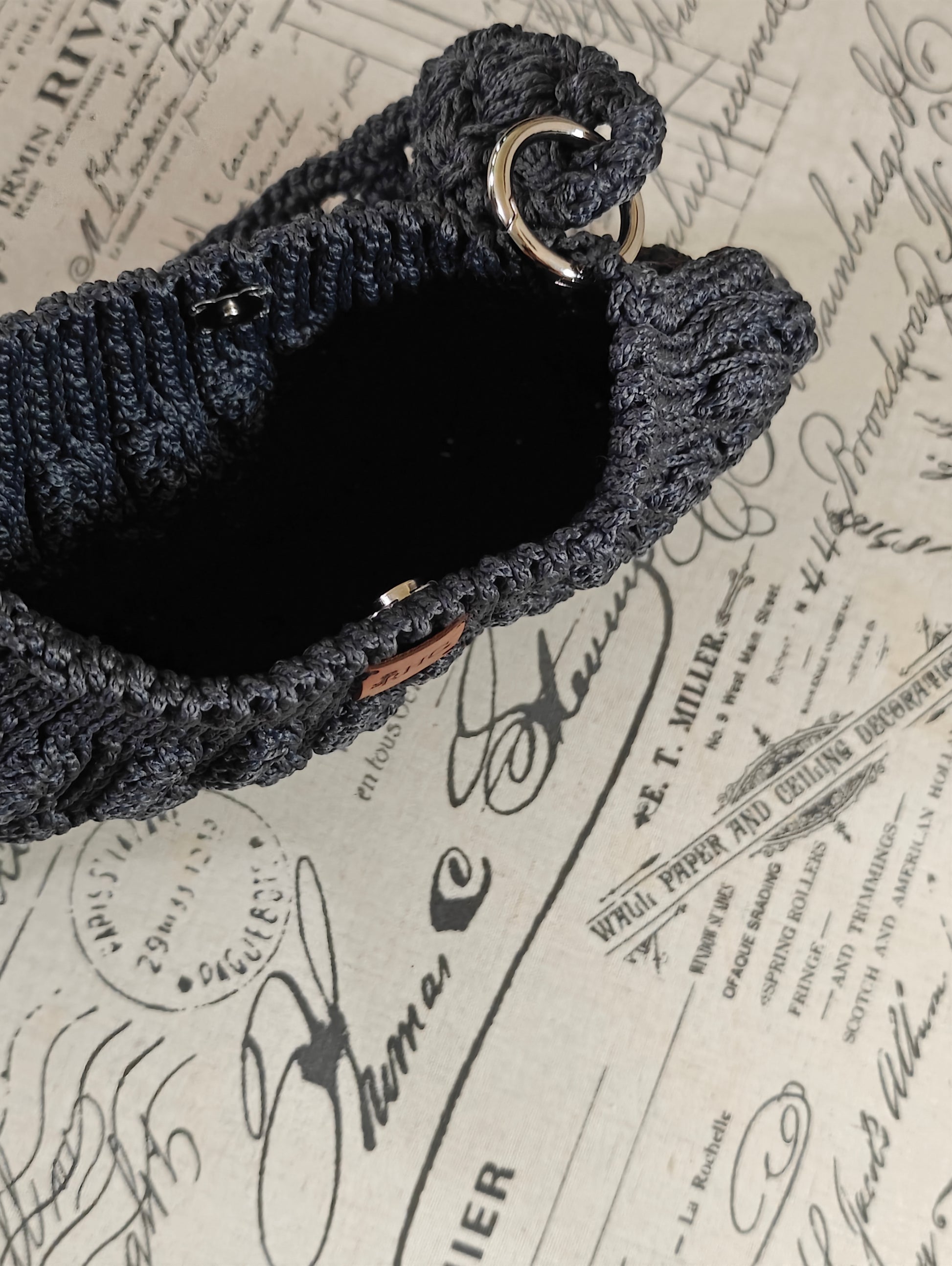 A close-up of a dark charcoal gray crocheted handbag with a wide shoulder strap, featuring a silver metal O-ring and a small brown leather brand tag, set against a vintage-style printed  background.