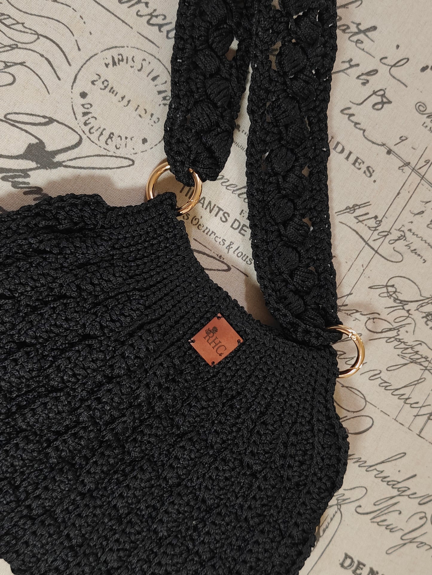 Close-up of a black crocheted shoulder bag with textured straps and gold-tone hardware, resting on a vintage-style script background. A small brown leather brand tag is visible on the bag.