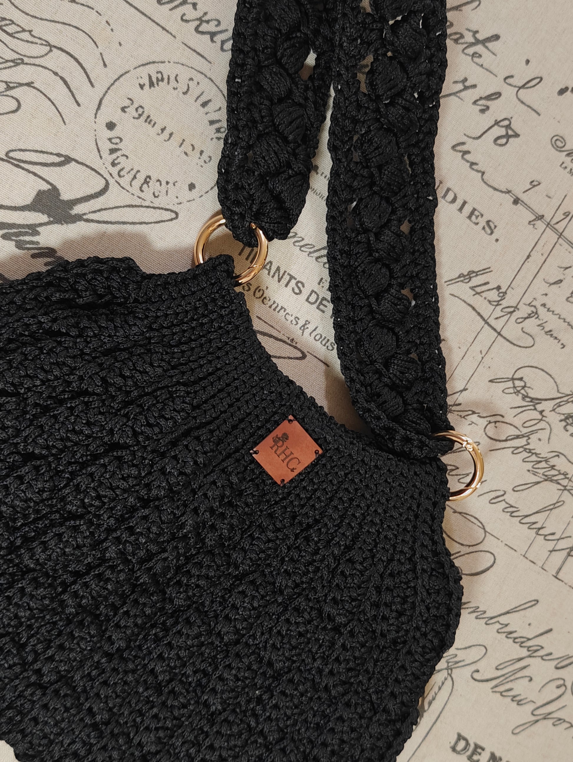 Close-up of a black crocheted shoulder bag with textured straps and gold-tone hardware, resting on a vintage-style script background. A small brown leather brand tag is visible on the bag.
